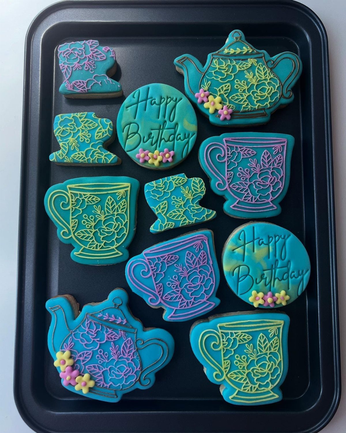 Decorated sugar cookies shaped like teapots and teacups arranged on a dark baking tray.