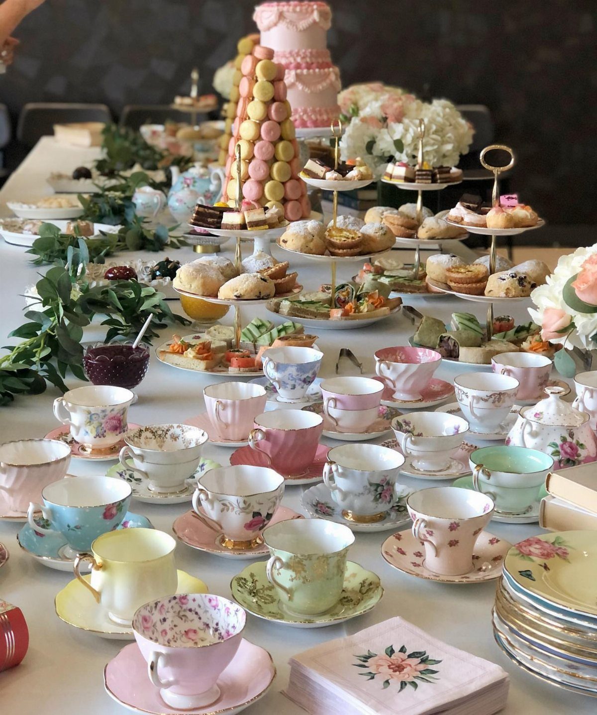 Mismatched vintage teacups and tiered dessert stands arranged on a long table for a tea party.
