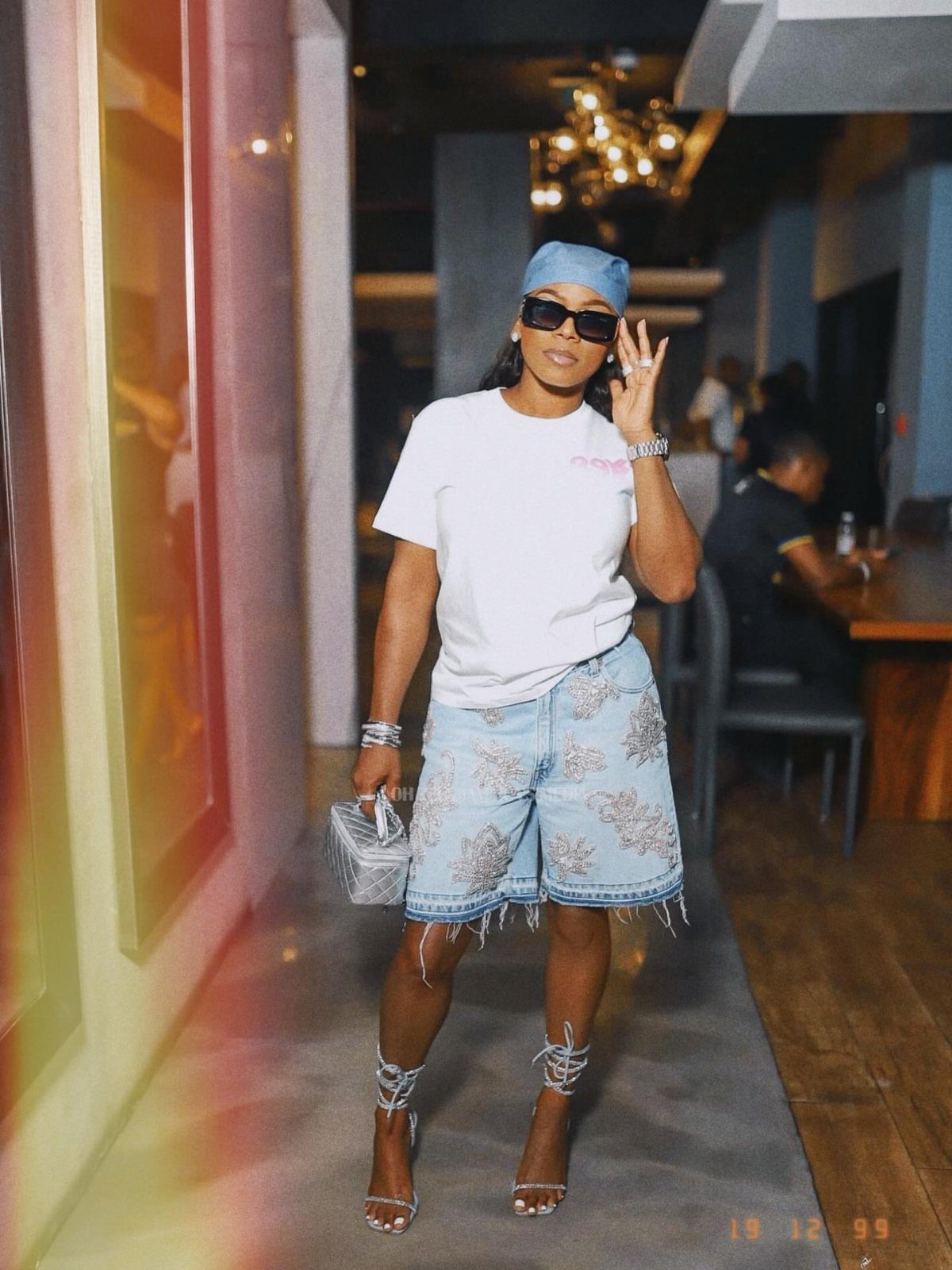 Woman posing in a white t-shirt, embroidered denim shorts, sunglasses, and silver strappy heels.