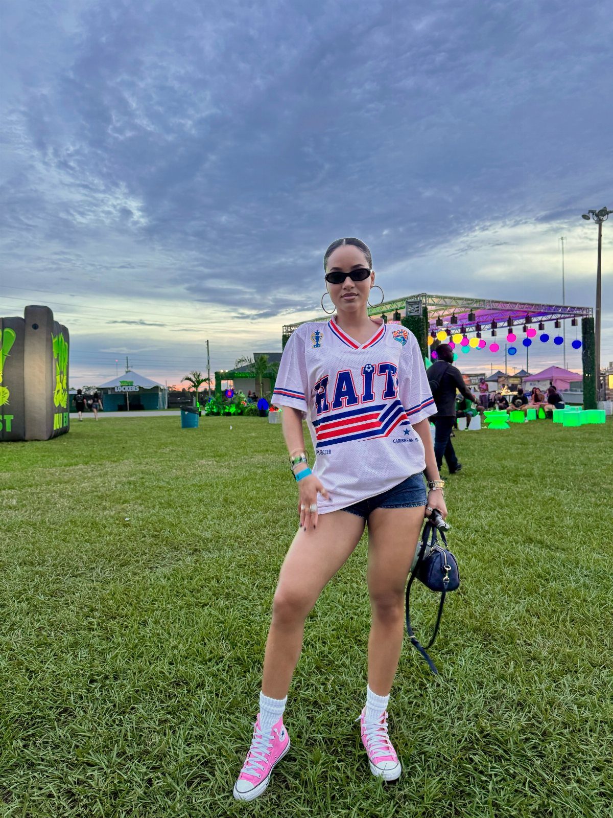 Woman in a white jersey, denim shorts, and pink sneakers standing on a grassy field.