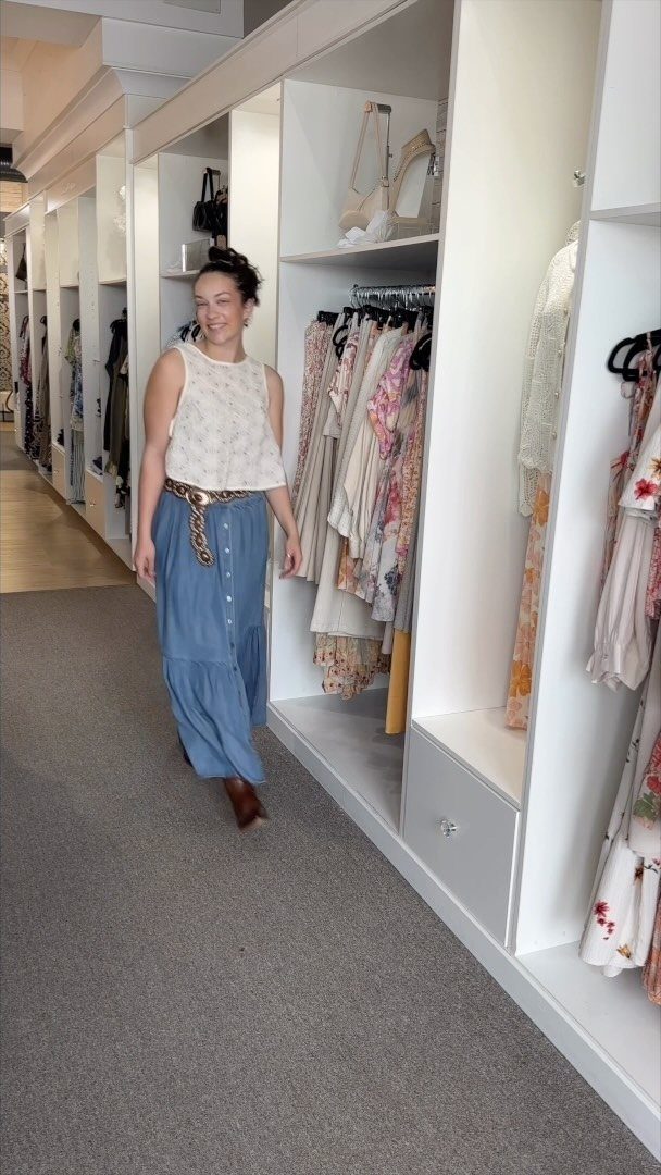 Woman walking in a clothing store wearing a white top and long blue skirt.
