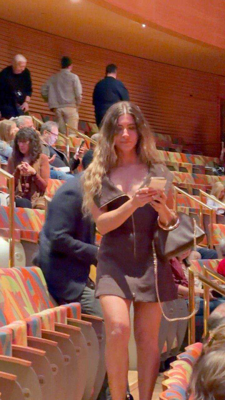 A woman in a dark brown mini dress stands in a theater with colorful seats.