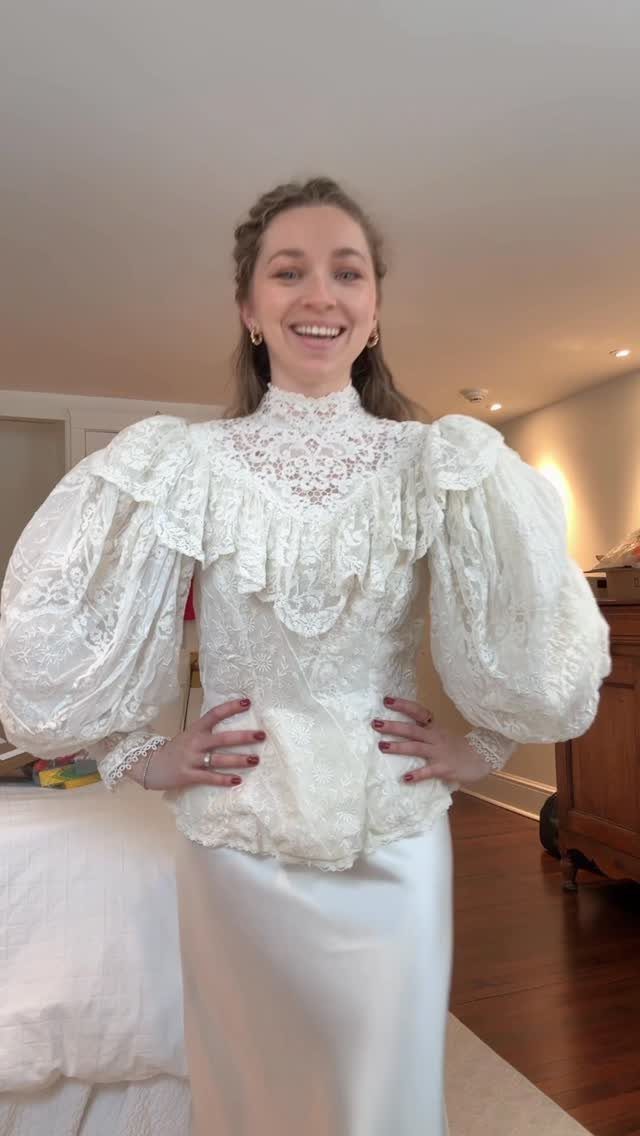 A smiling woman wearing a white lace ruffled blouse and a long white satin skirt.