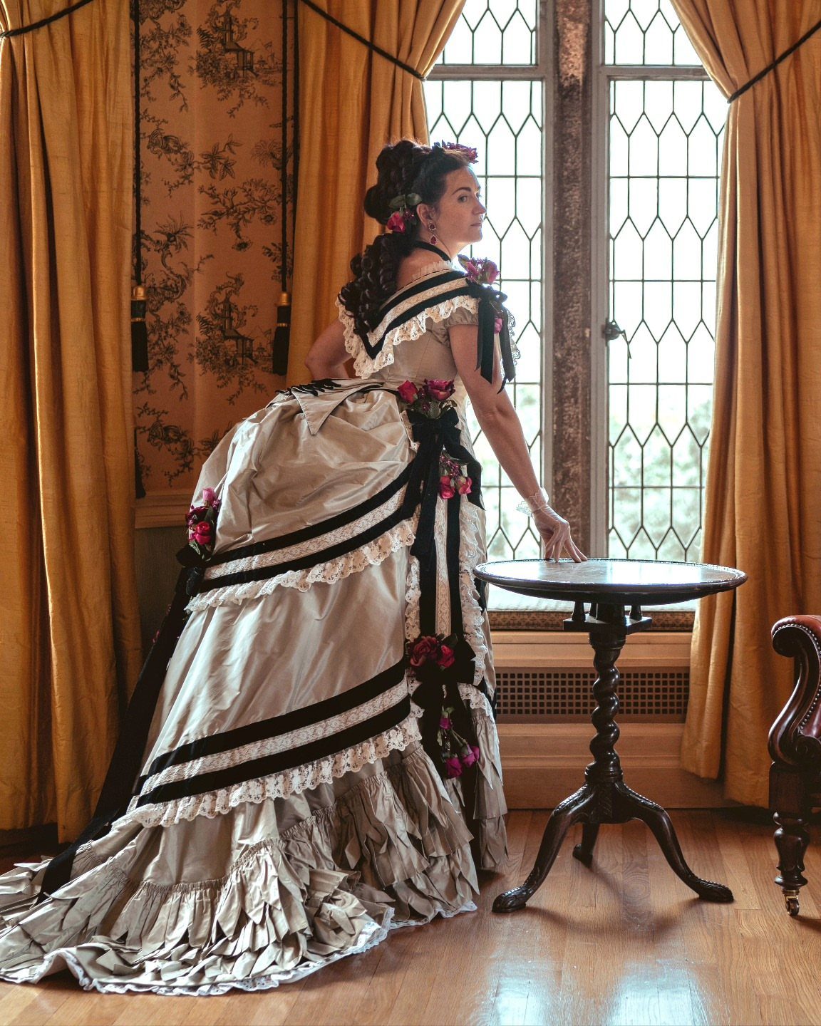 A woman wearing a Victorian bustle period dress standing next to a small table by a window.