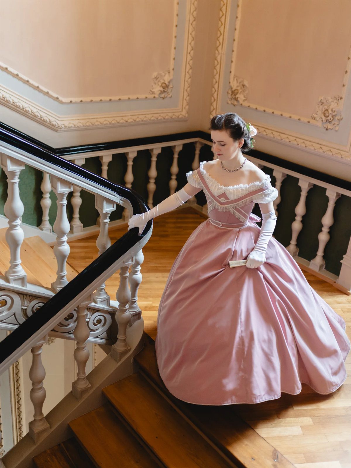 A woman in a wide pink Victorian gown walking down an ornate wooden staircase.