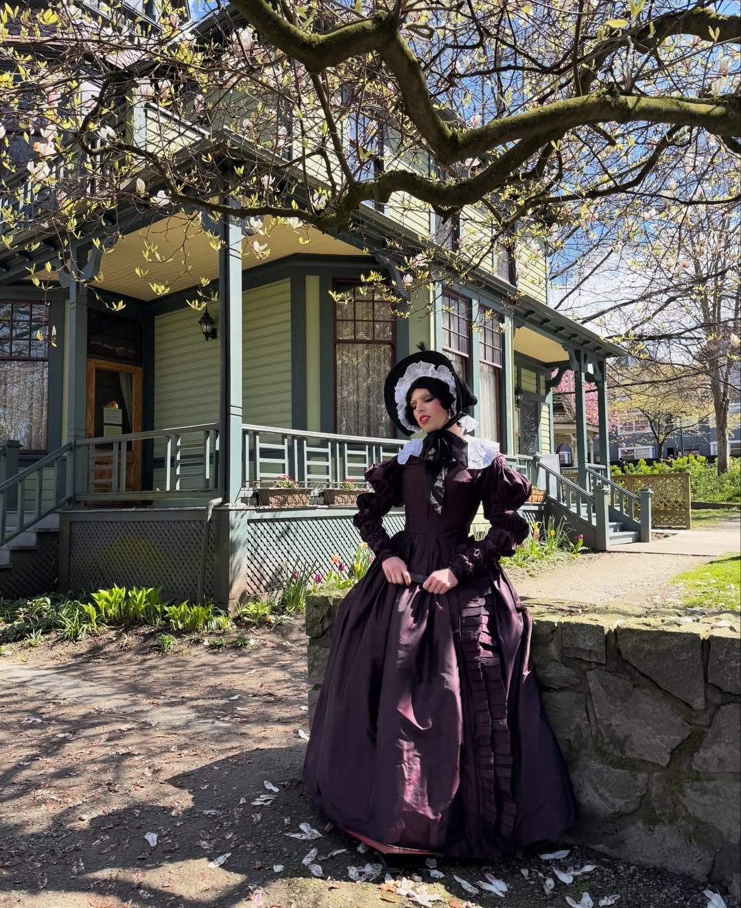 A woman wearing a dark purple Victorian day dress and black bonnet stands near a house.