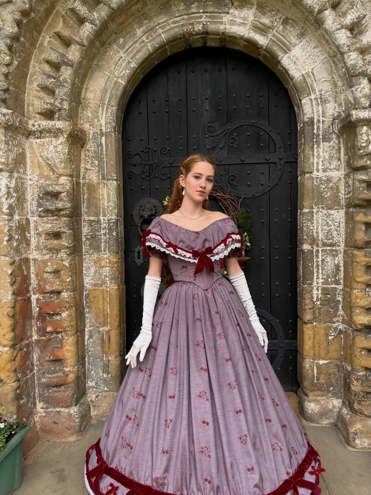 A woman in a lavender Victorian ball gown with red trim standing before an arched stone doorway.