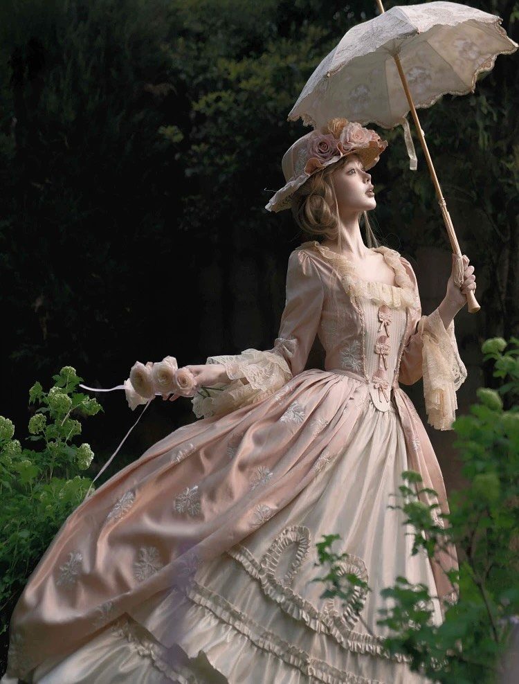 A woman wearing a pink Victorian gown and floral hat holding a lace parasol in a garden.