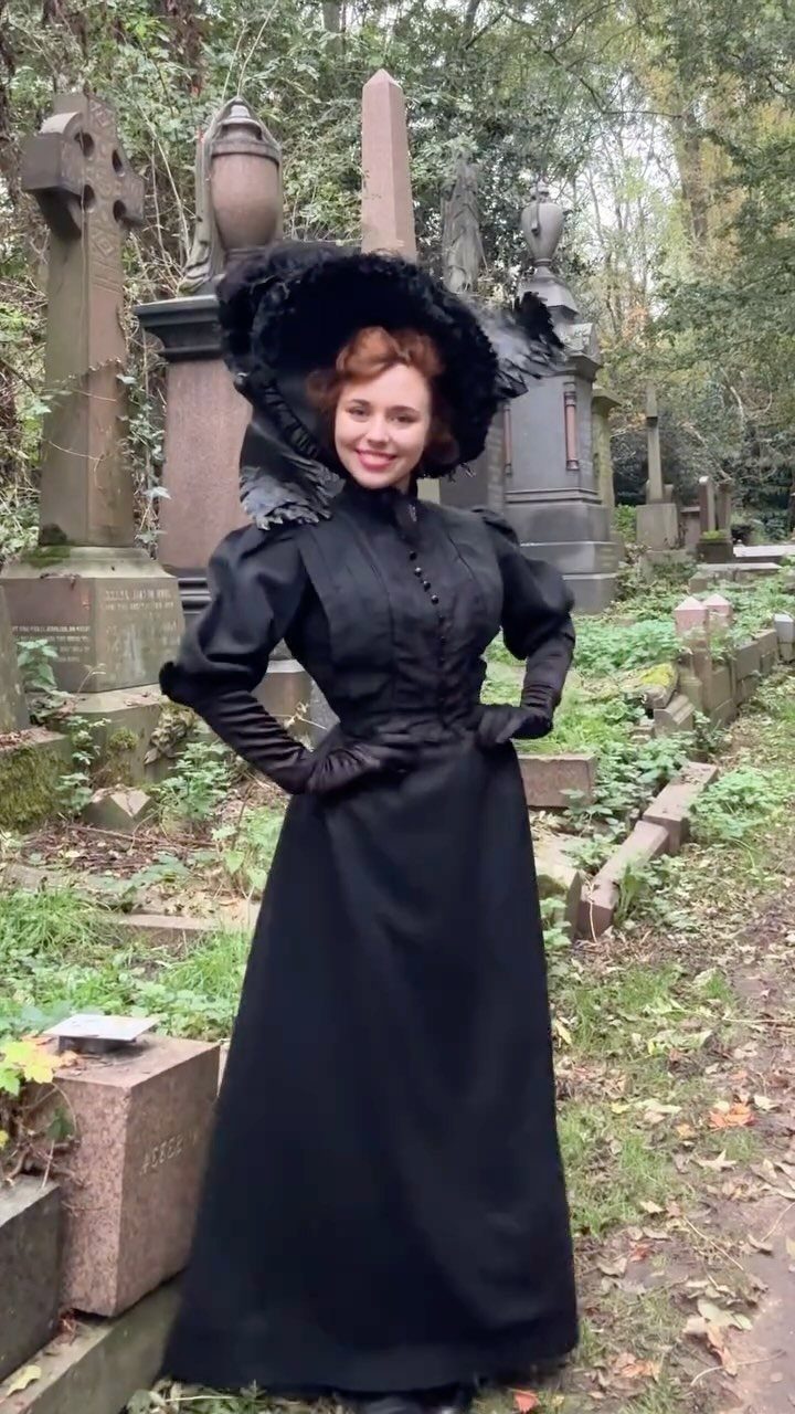 A woman wearing a black Victorian-inspired dress and large hat posing in a cemetery.
