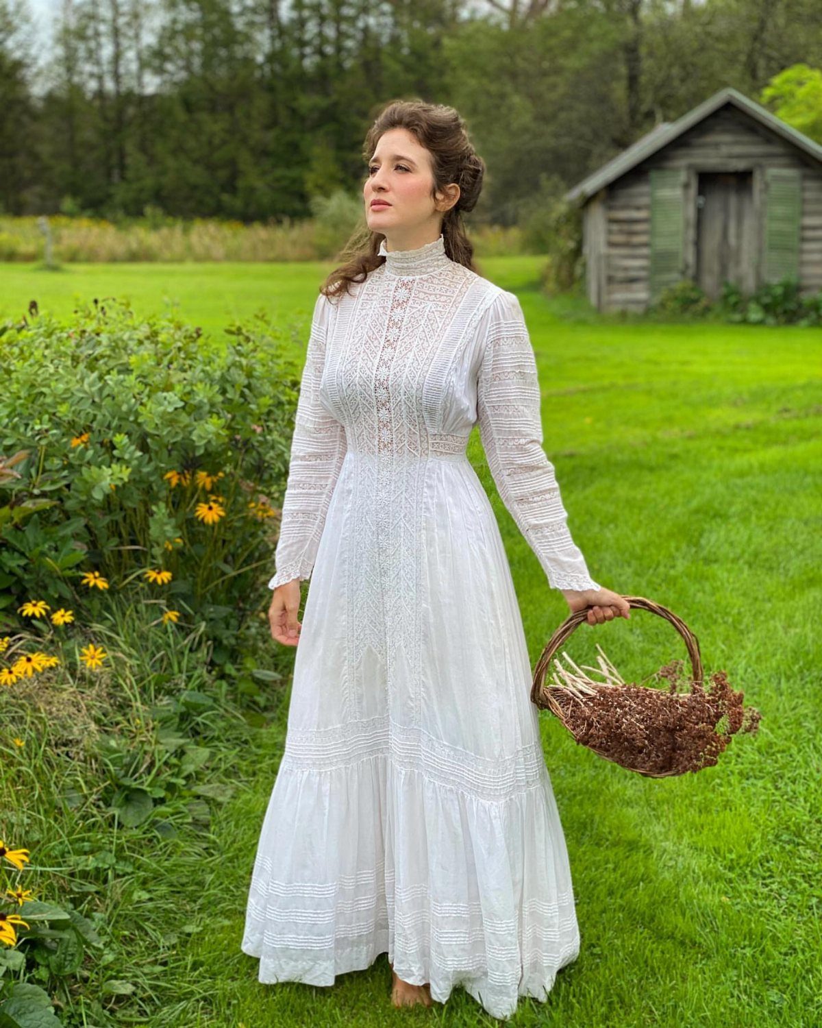 Woman in a long white lace Edwardian tea dress standing in a green field holding a basket.