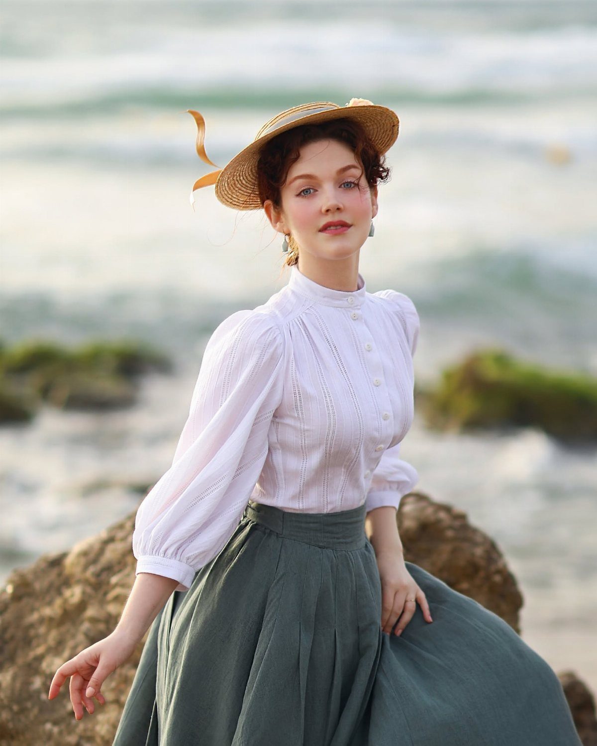 A woman in a white vintage blouse and grey skirt posing by the ocean.