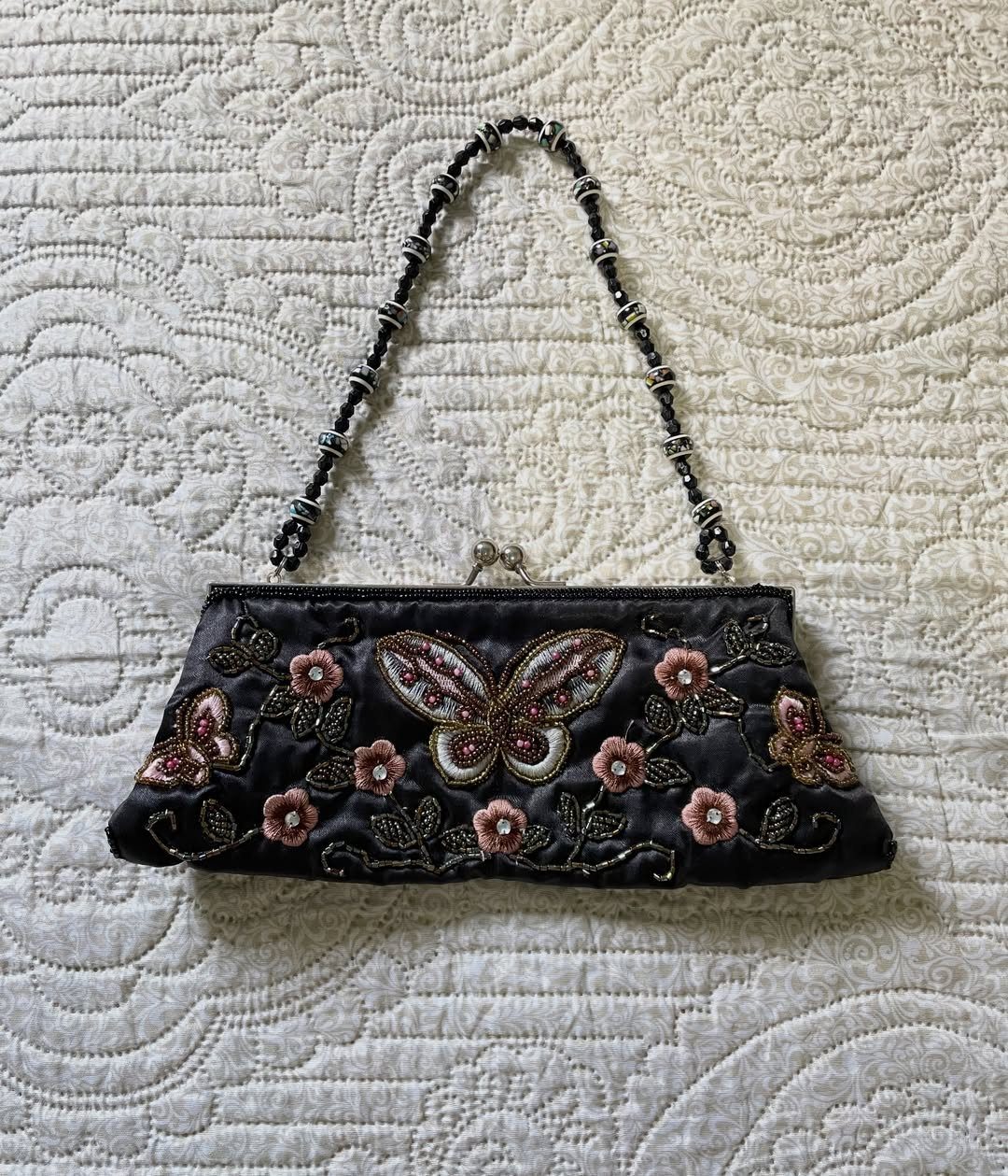 Embroidered black satin clutch bag with butterfly and floral patterns on white quilted fabric.