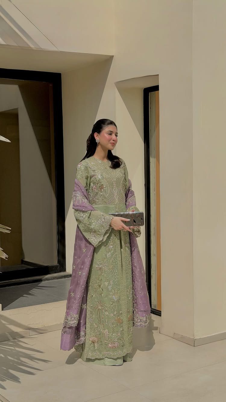 A woman in a light green embroidered dress and lavender shawl standing by a building.