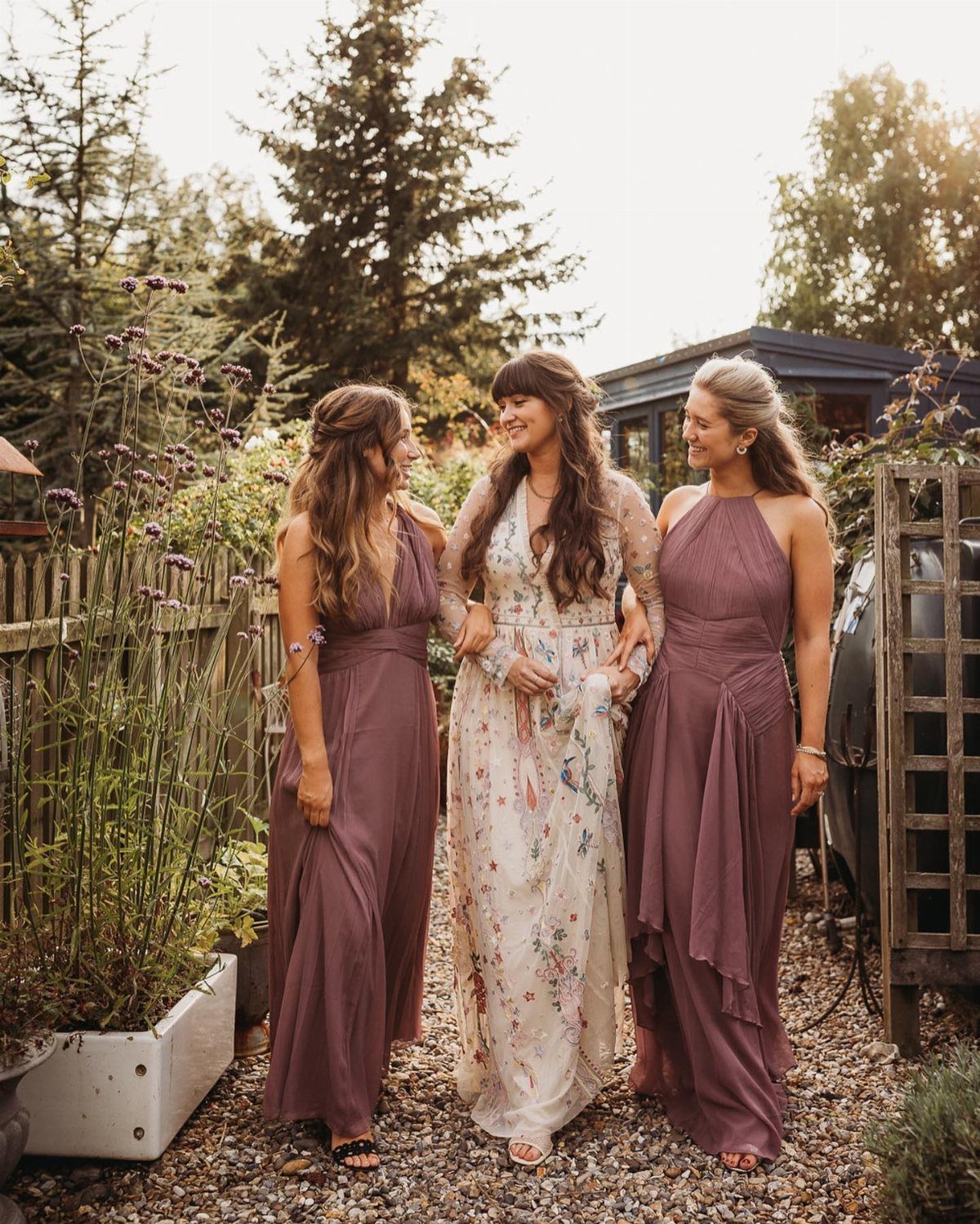 Three women walking through a garden wearing elegant midi-length floral and mauve dresses.