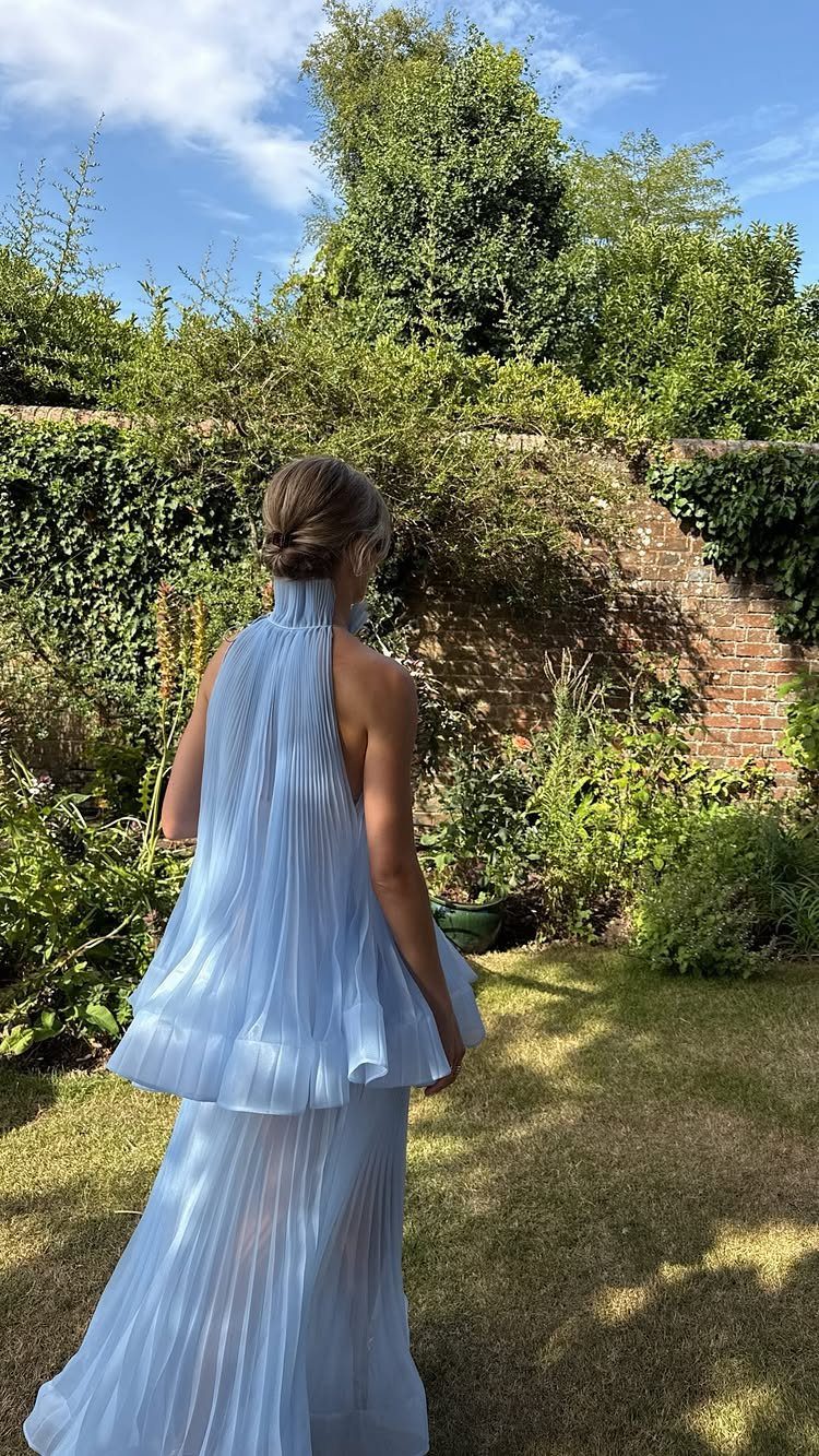 Woman in a light blue pleated halter gown standing in a lush green garden.