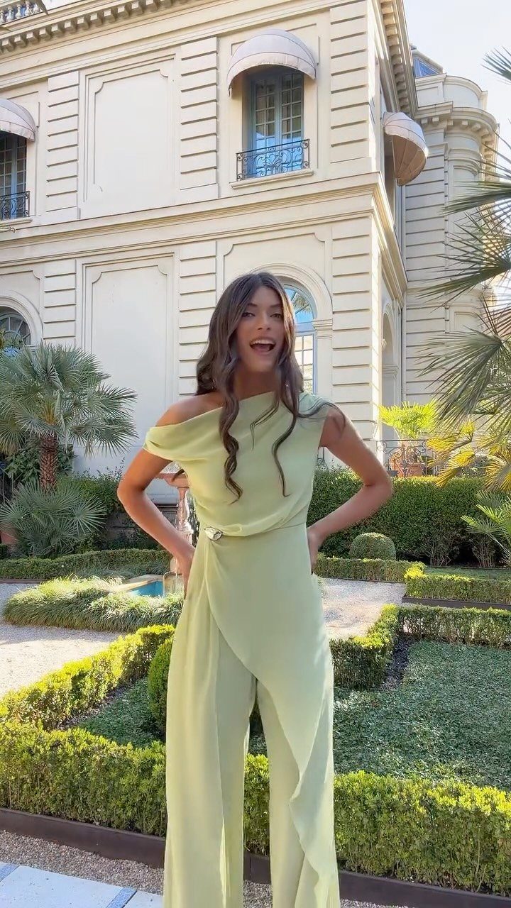 A woman wearing a light green one-shoulder jumpsuit standing in a manicured garden.