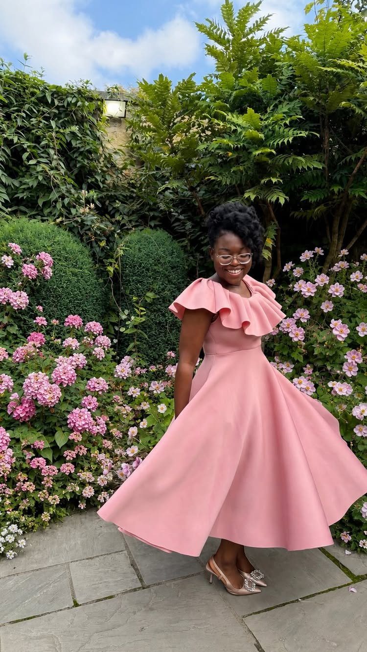 A woman in a pink ruffled midi dress posing in a lush garden with pink flowers.