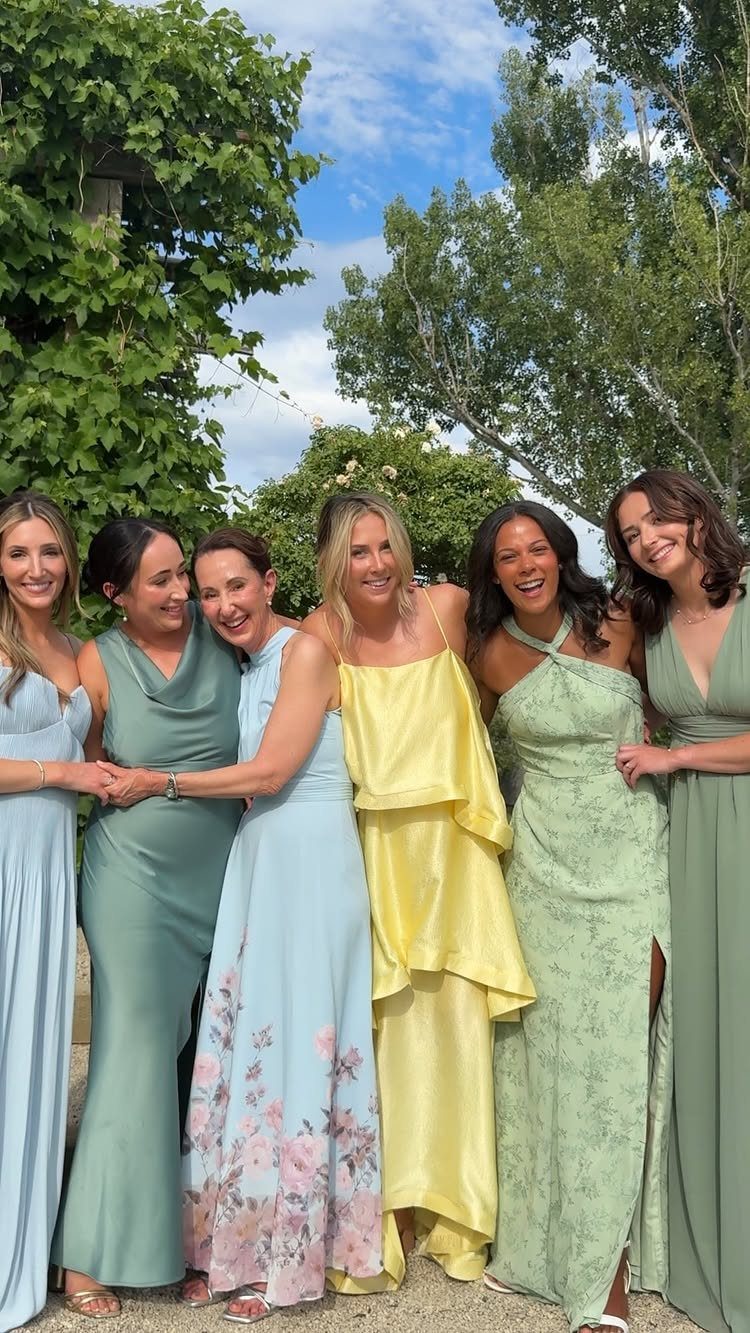 Six women in elegant pastel summer dresses posing together in a lush green garden.