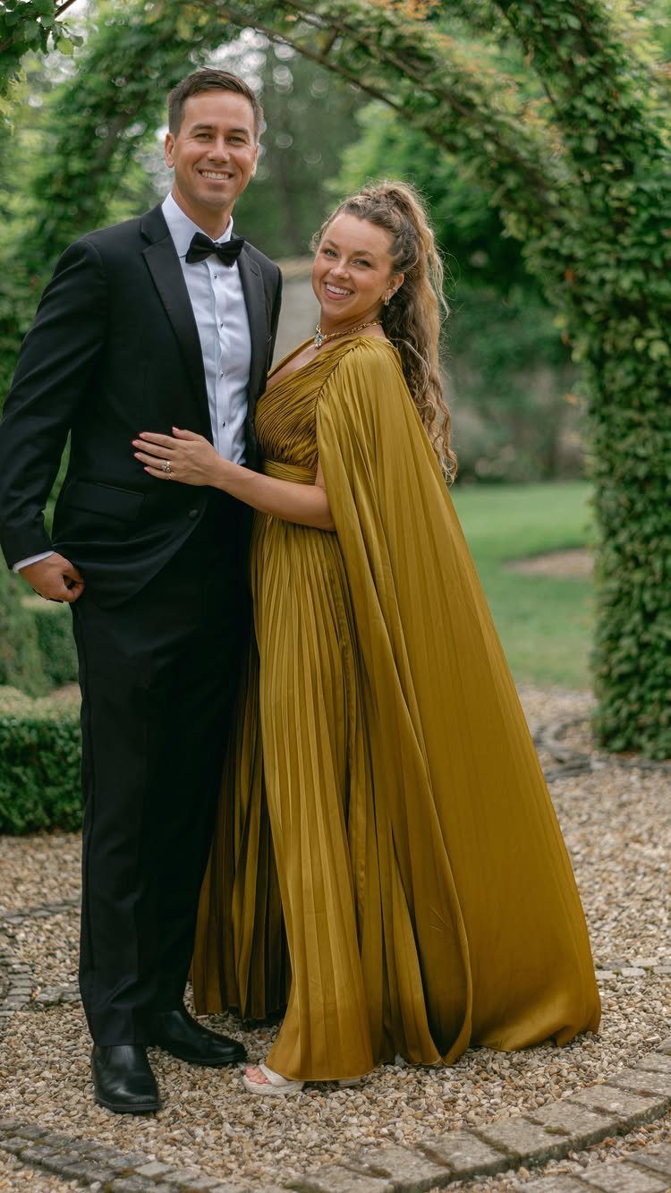 A smiling couple posing in a garden, the woman wearing a long pleated mustard-yellow dress.