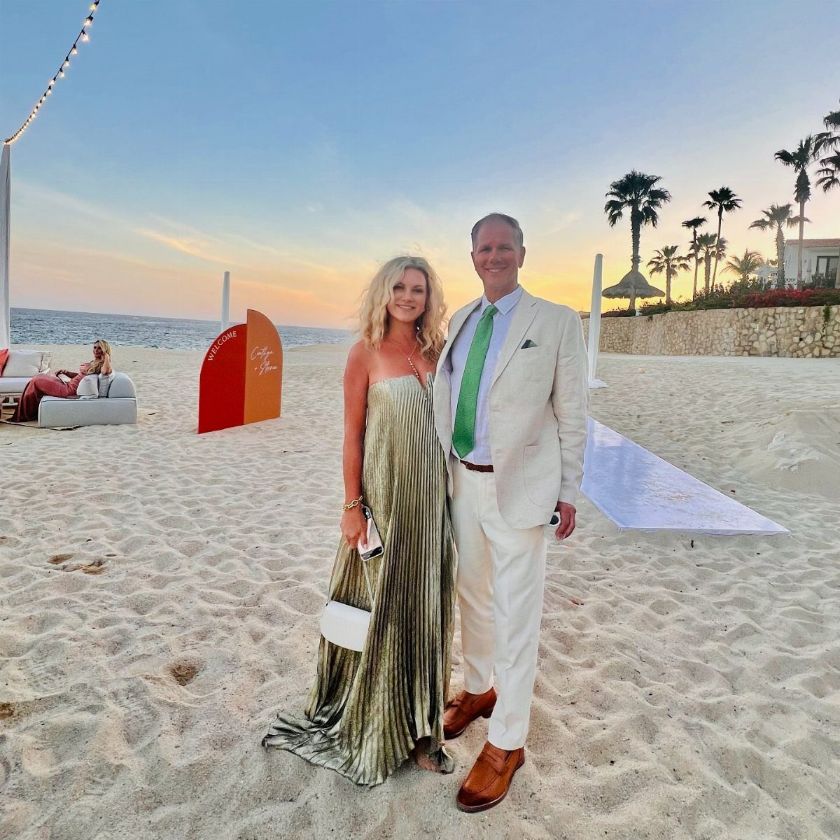 A man in a beige suit and a woman in a gold pleated dress standing on a beach.