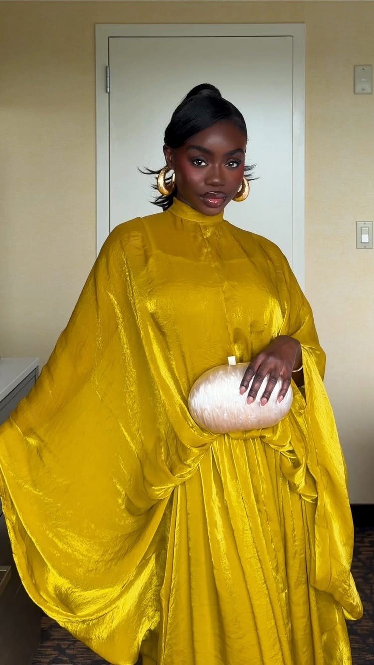 A woman in a flowing mustard-yellow satin gown holding a white clutch bag.