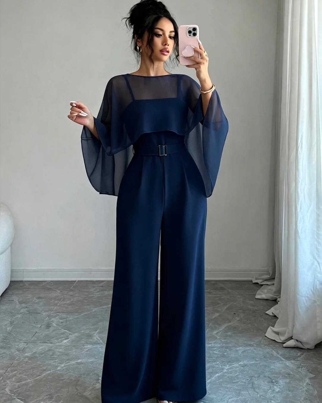 Woman in a navy blue wide-leg jumpsuit with sheer cape sleeves taking a mirror selfie.