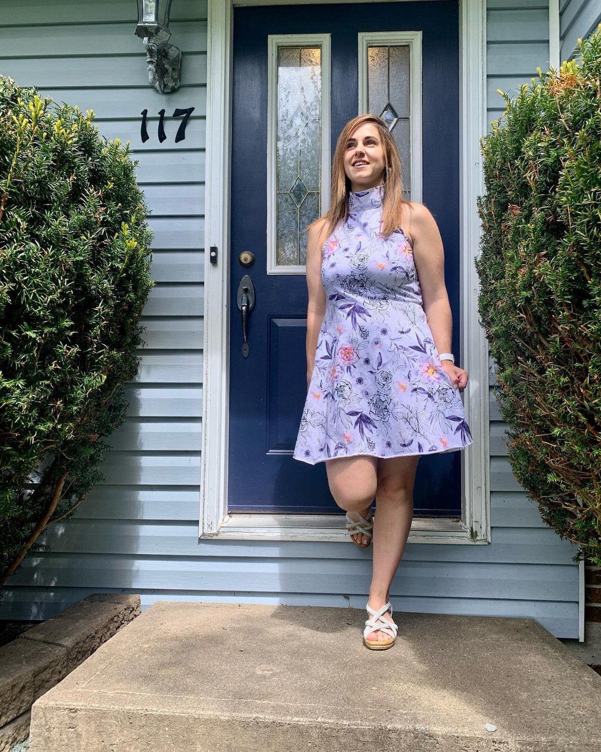 Woman in a light purple floral sundress and white sandals standing on a porch.