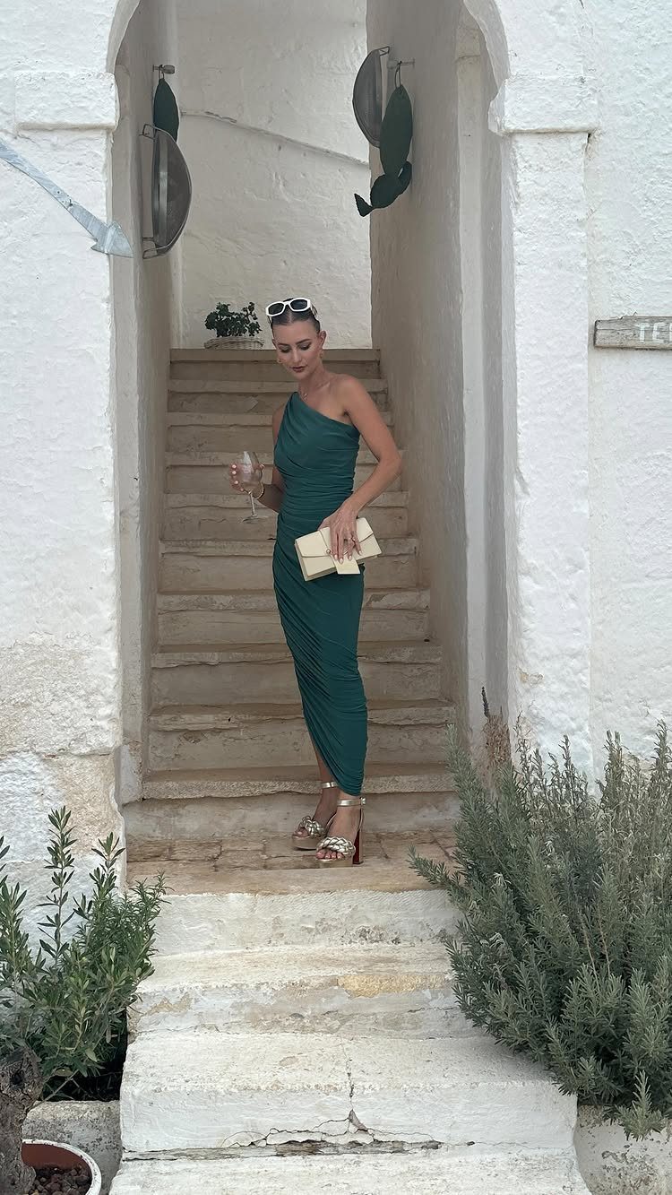 Woman in a dark green one-shoulder dress and gold heels posing on white stone stairs.