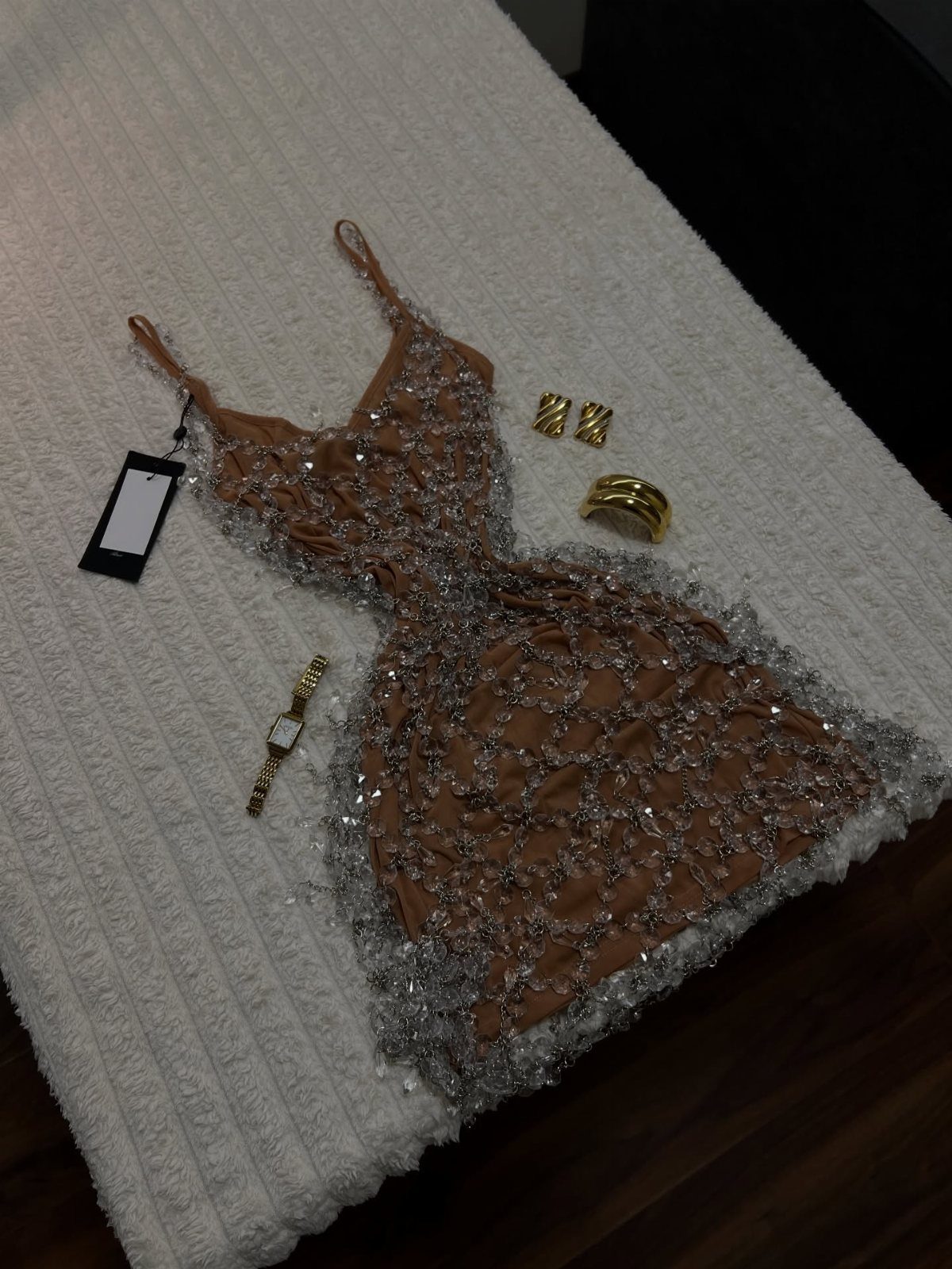 A tan sequined mini dress laid out with gold jewelry on a white textured bedspread.