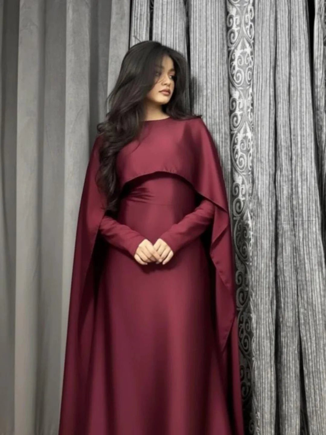 Woman wearing a long-sleeved burgundy satin dress with a dramatic integrated cape.