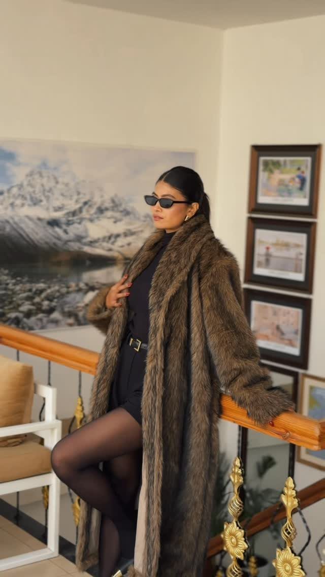 Woman in a long brown faux fur coat and black dress posing indoors.
