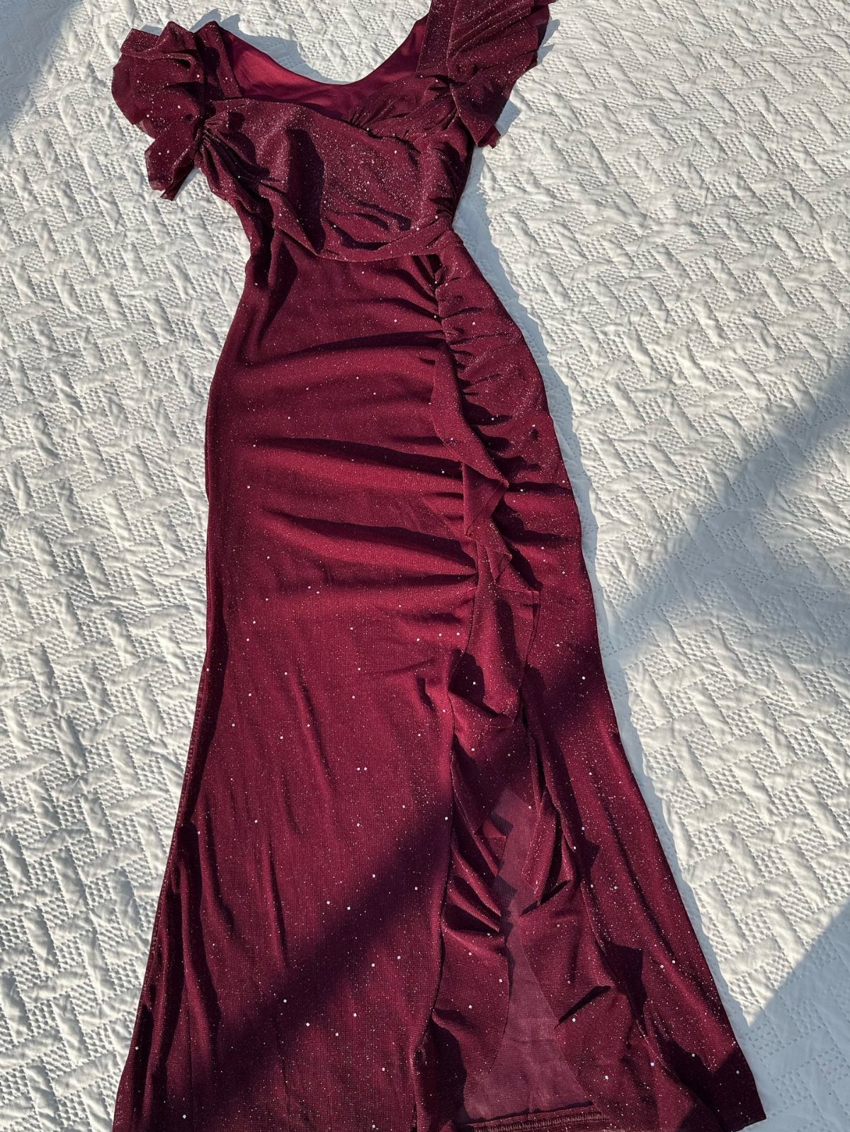 A shimmering burgundy maxi dress with ruffles lying on a white textured fabric surface.