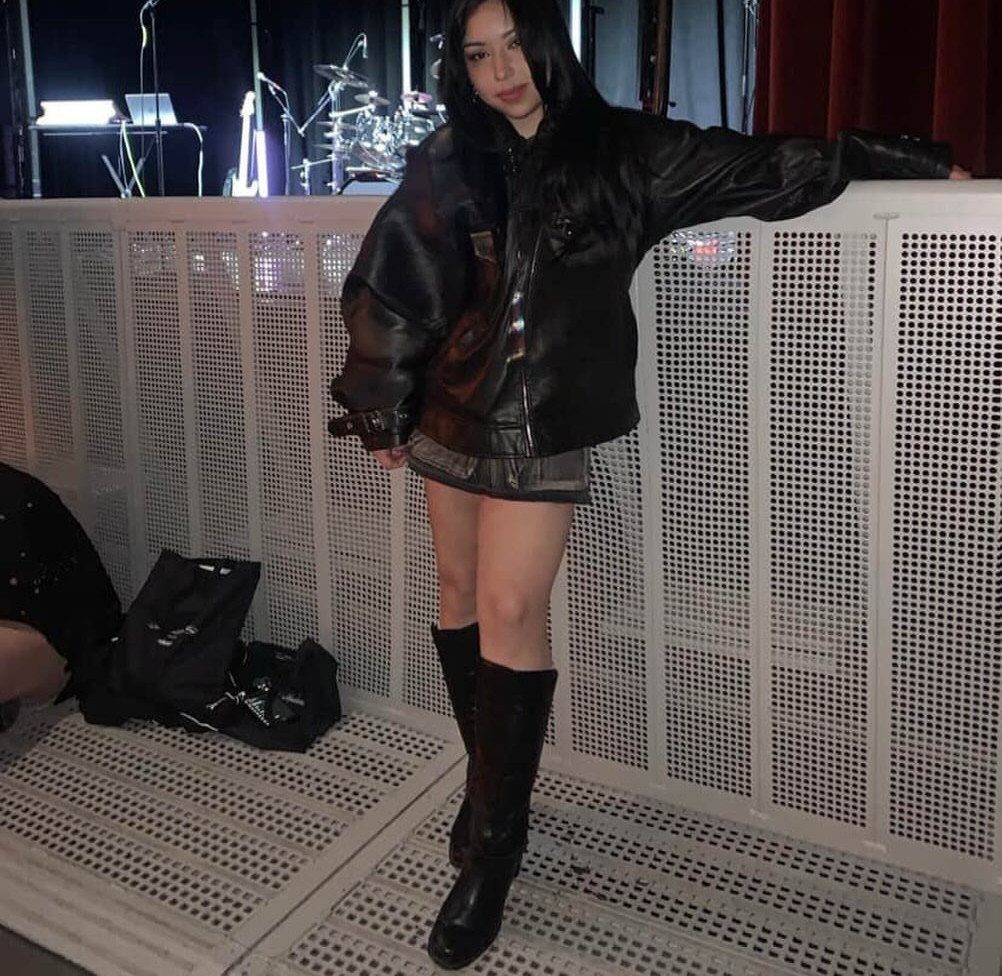 Woman in a black leather jacket, skirt, and tall boots standing near a stage.