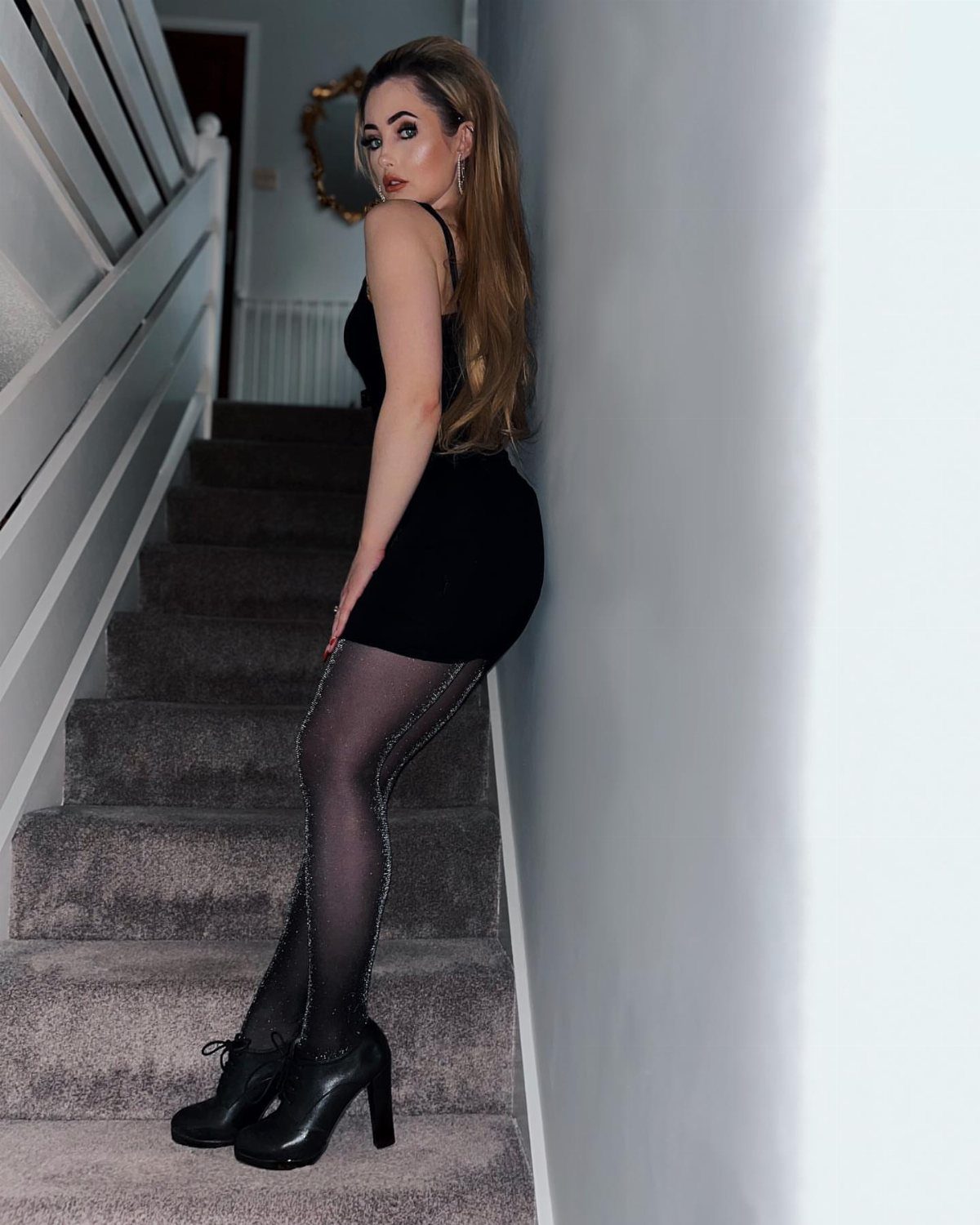 Woman in a black mini dress, shimmering tights, and high-heeled boots posing on stairs.
