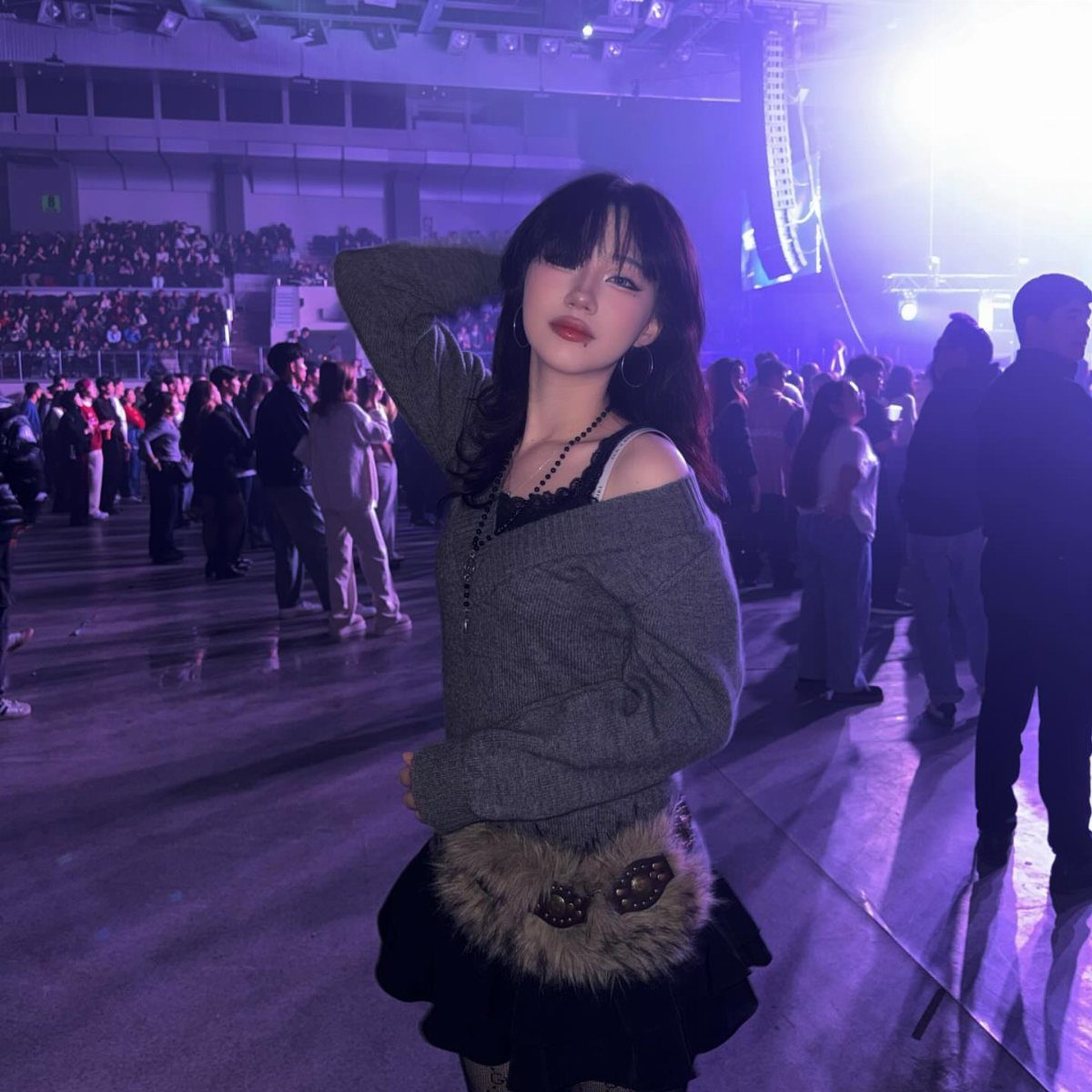 Young woman posing in a grey sweater and fur-trimmed skirt at a concert.