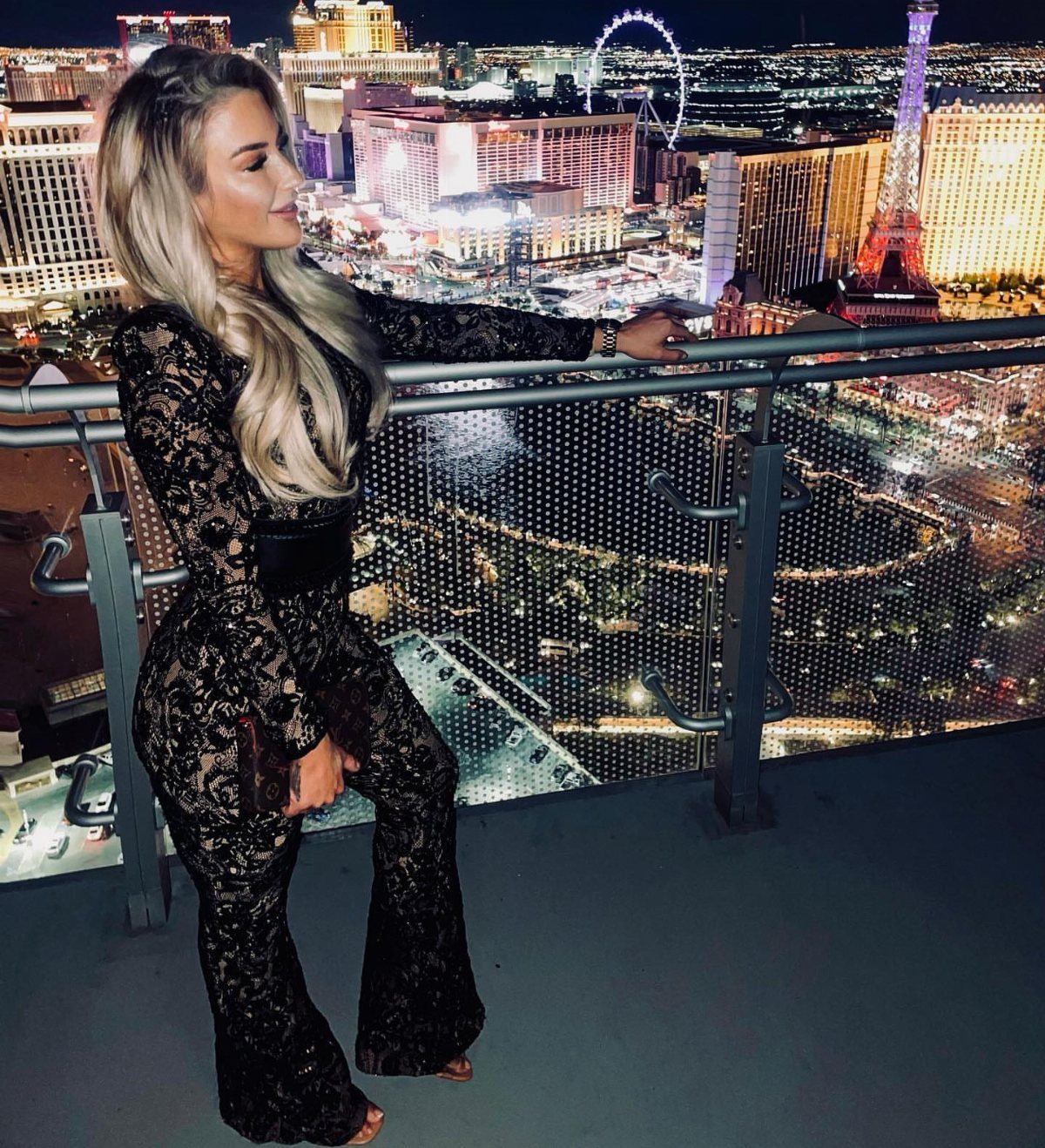 Blonde woman in a black lace jumpsuit posing on a balcony overlooking the Las Vegas night skyline.