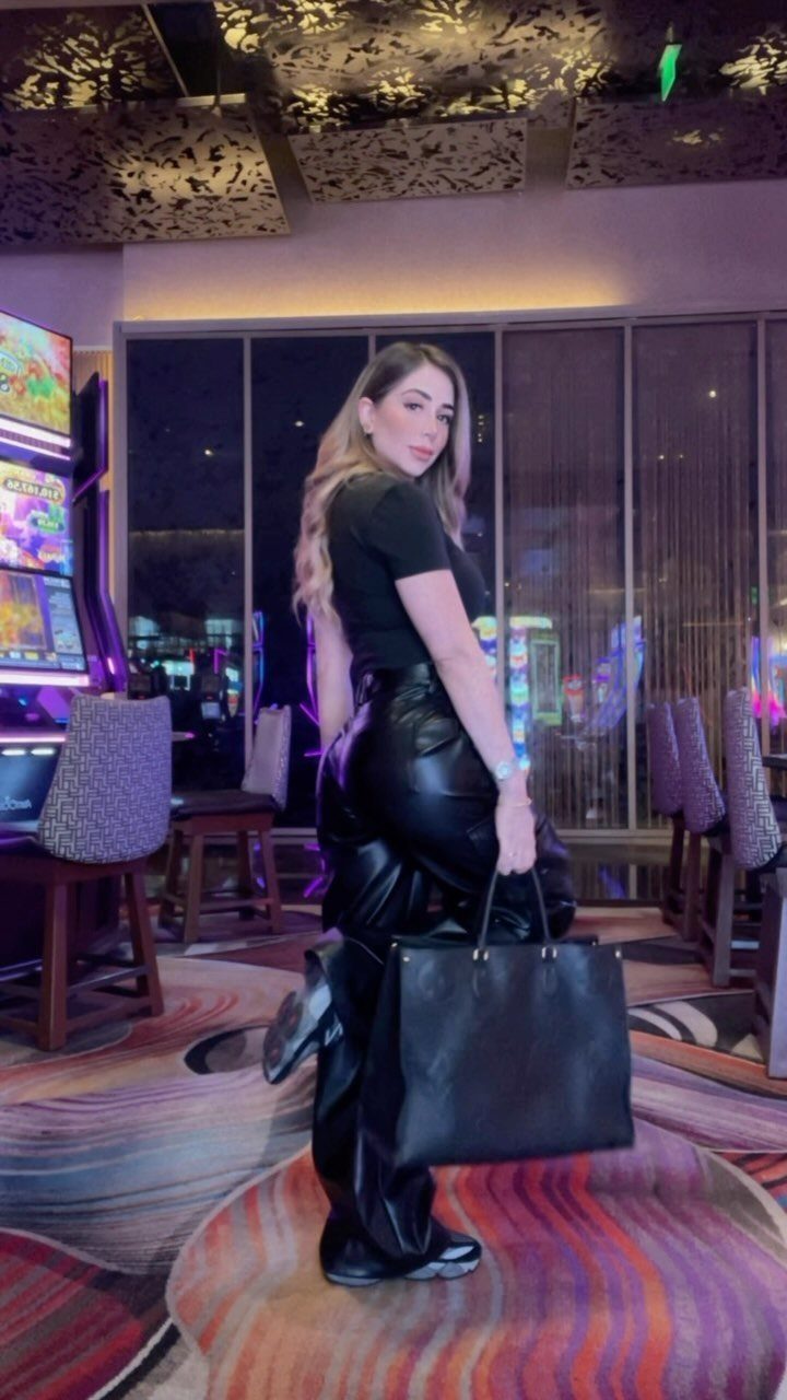 Woman in black leather pants and t-shirt posing in a casino setting with a large tote bag.