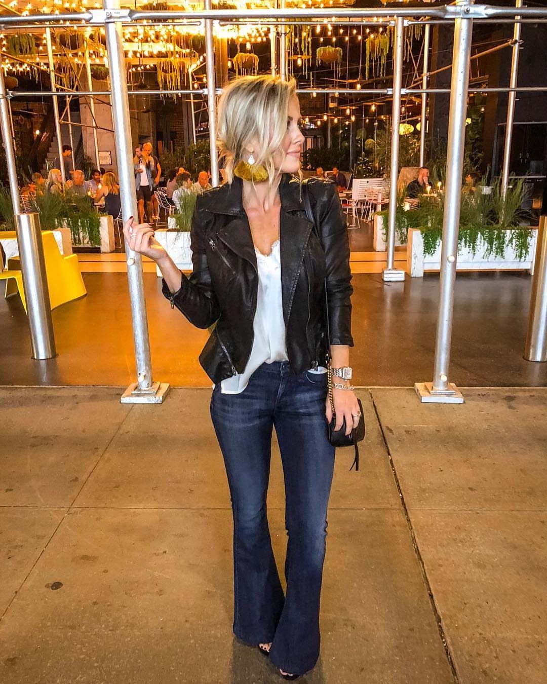 Blonde woman wearing a black leather jacket, white top, and flared blue jeans standing outdoors at night.