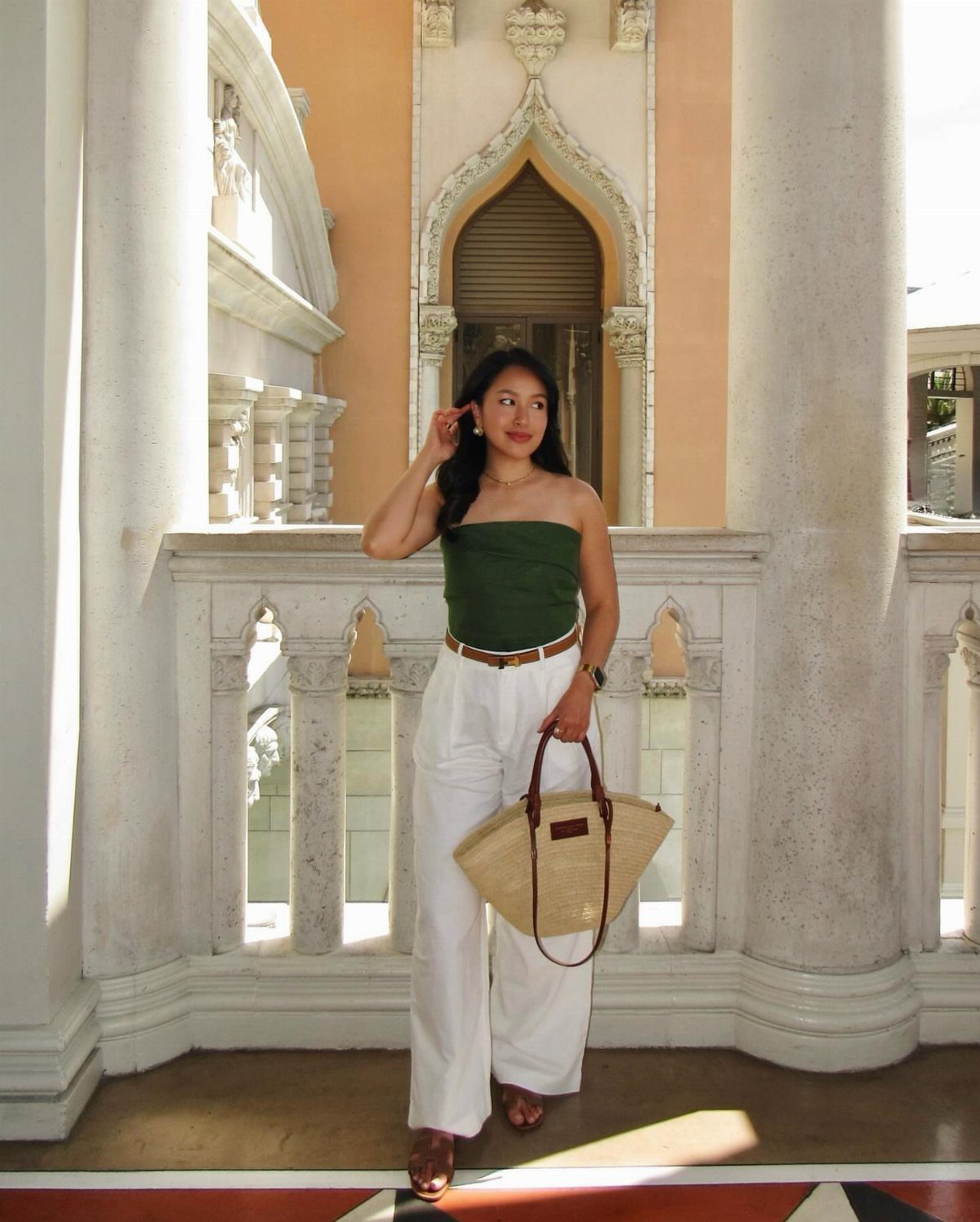 Woman in green strapless top and white wide-leg trousers holding a straw bag on a balcony.