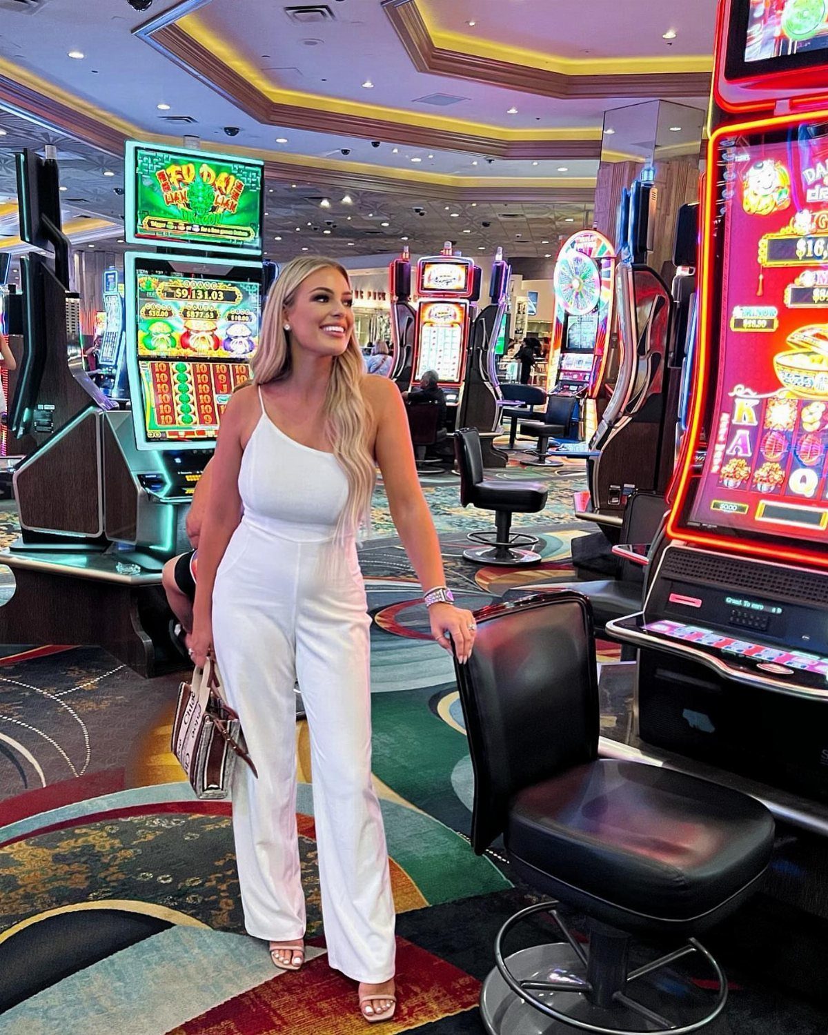 A blonde woman wearing a white jumpsuit stands smiling in a brightly lit casino environment.