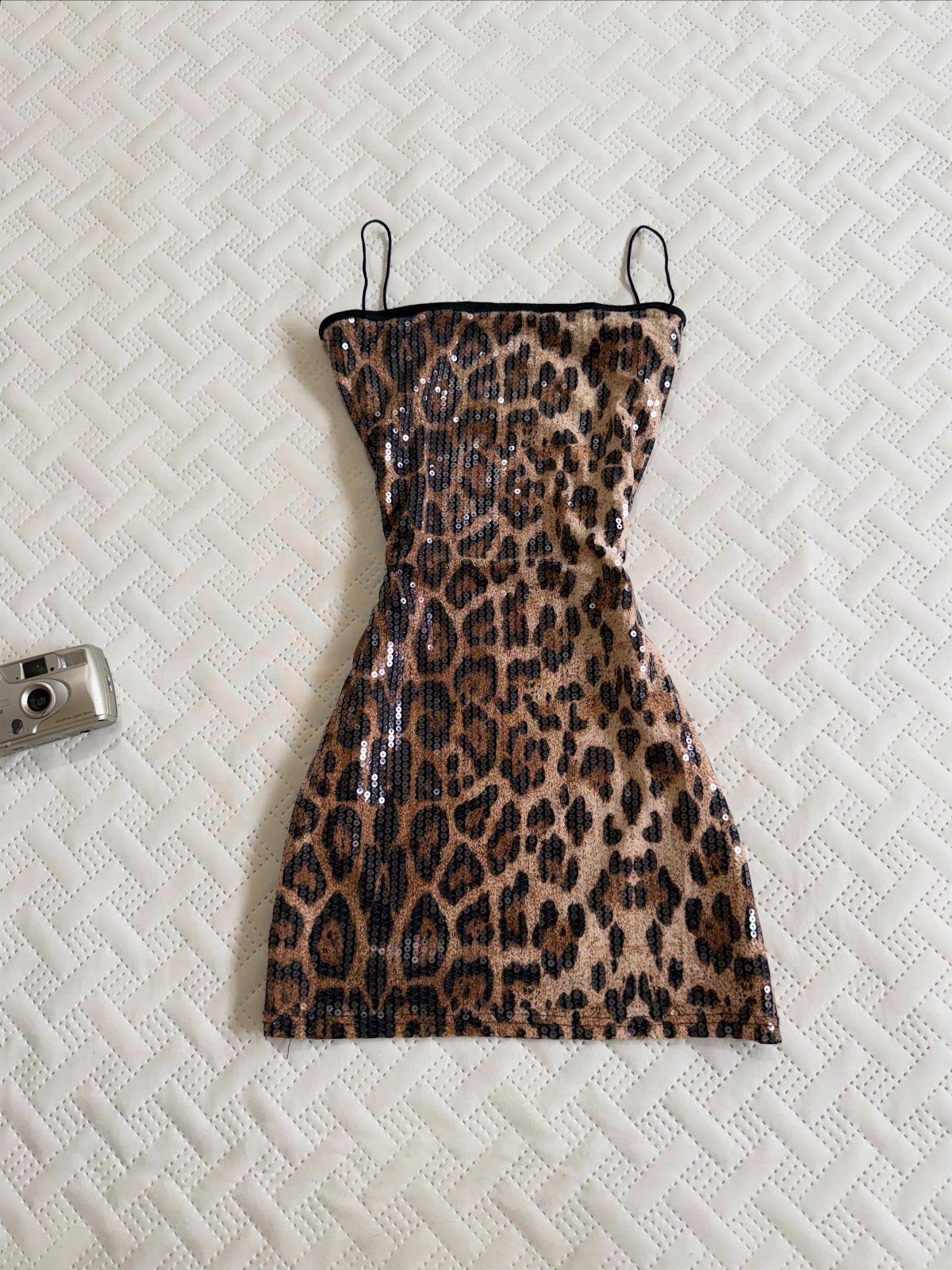 Leopard print sequined mini dress with thin straps lying on a white textured background.