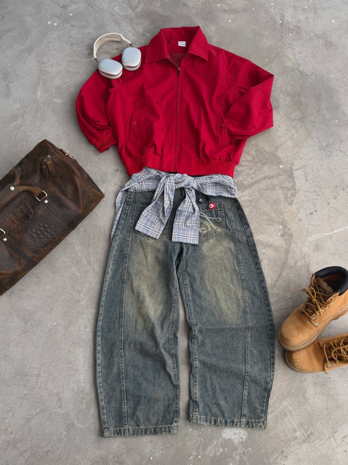Red jacket, baggy jeans, plaid shirt, headphones, brown bag, and tan boots laid on concrete.