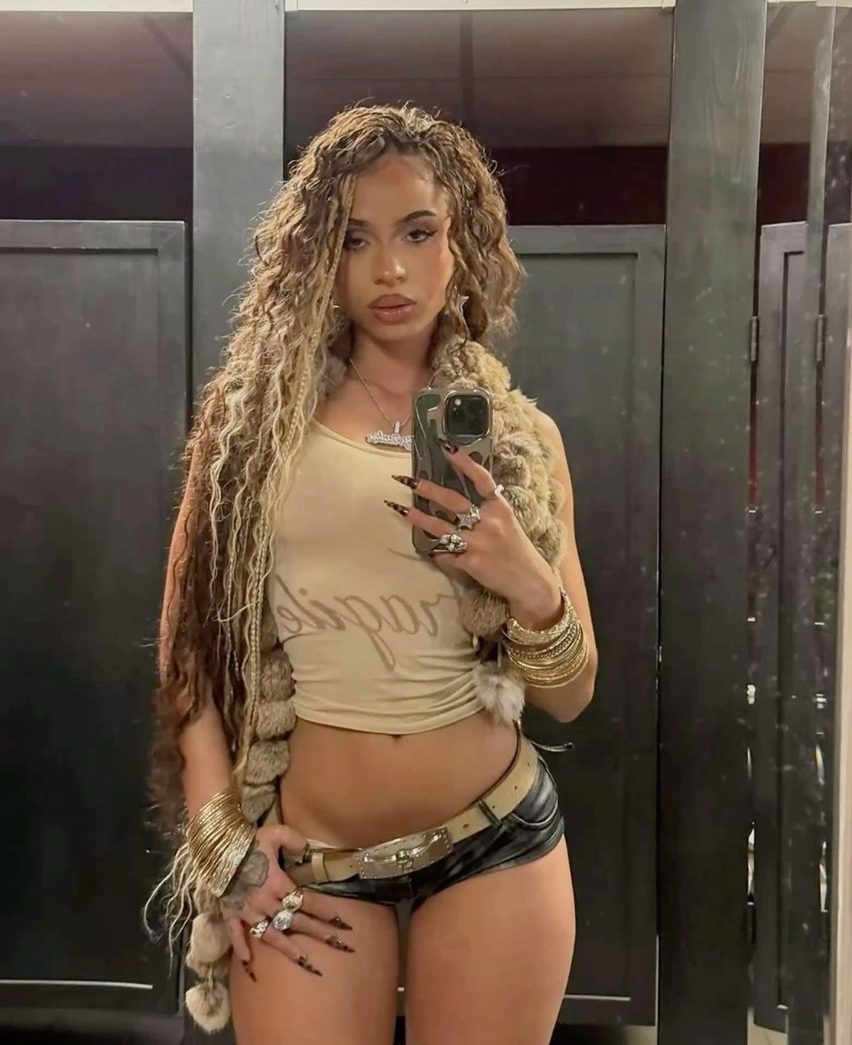 Woman with long curly hair posing in a tan crop top and black leather shorts.