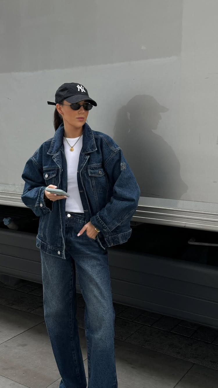 Woman posing in a dark denim jacket and matching denim jeans with a black cap.