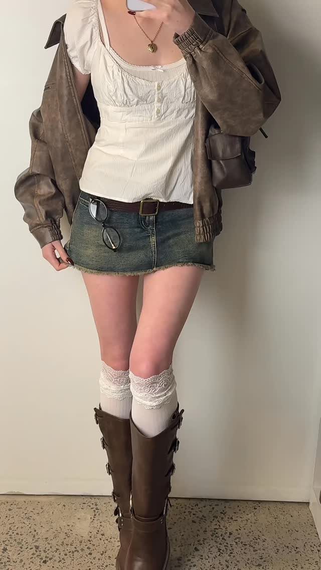 A person posing in a white top, denim mini skirt, brown jacket, and tall brown boots.
