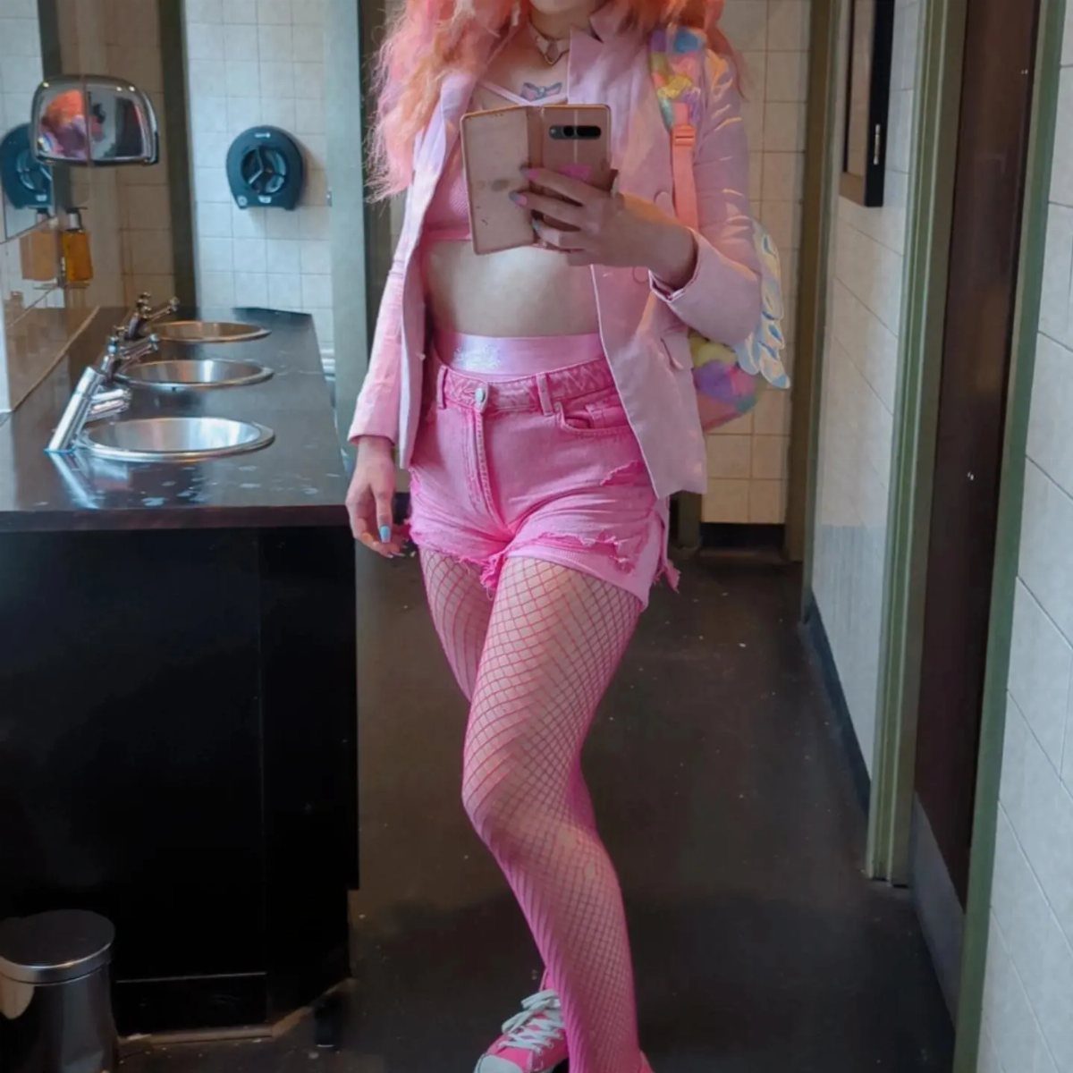 Person with pink hair wearing a pink outfit and fishnet tights posing in a restroom mirror.