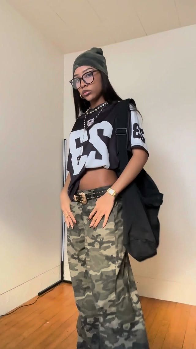 A woman posing in a cropped jersey, camouflage pants, and a green beanie against a white wall.