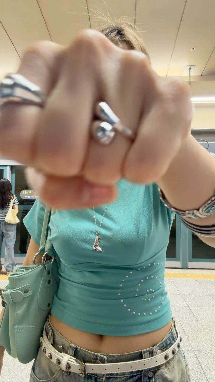 Person wearing a teal cropped t-shirt, studded belt, and chunky silver rings in a public space.
