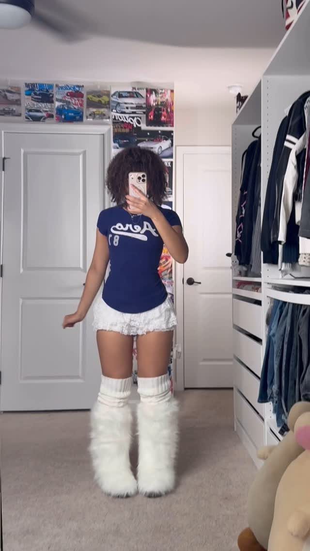 Person wearing a blue t-shirt, white shorts, and large white fuzzy leg warmers posing in a bedroom.