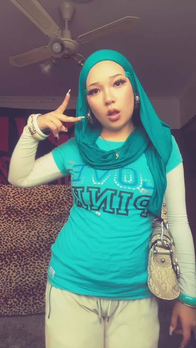 Young woman in teal hijab and pink graphic t-shirt posing with a peace sign.